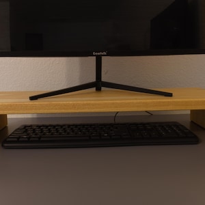 May include: A light-colored wooden monitor stand with a black keyboard underneath. A black computer monitor sits on top of the stand. The stand is rectangular with a raised platform.