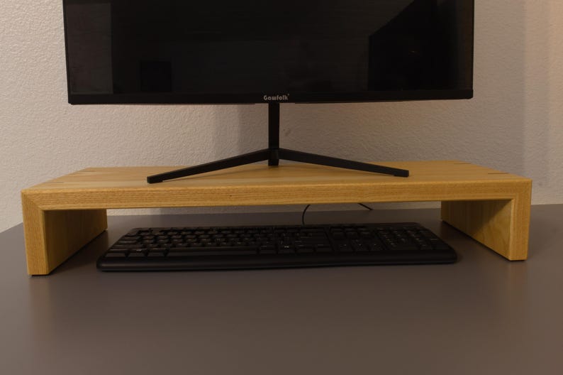 May include: A light-colored wooden monitor stand with a black keyboard underneath. A black computer monitor sits on top of the stand. The stand is rectangular with a raised platform.