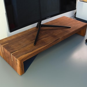 Puede incluir: Un soporte de monitor de madera con un acabado marrón intenso. El soporte tiene forma rectangular con una plataforma elevada y una parte inferior azul oscuro. Se ven detalles dorados en las esquinas. Un monitor de ordenador negro está encima del soporte.