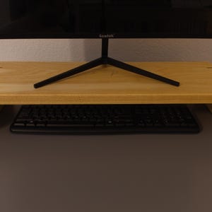 May include: A light-colored wooden monitor stand with a rectangular design. The stand has a keyboard underneath and supports a black computer monitor. The stand is made of light-colored wood and has a simple, modern aesthetic.