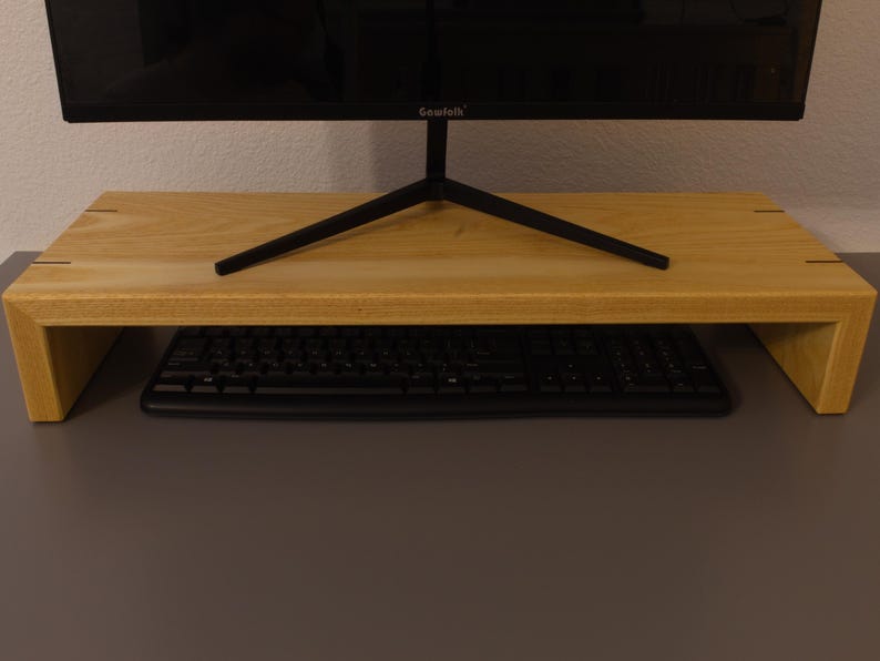 May include: A light-colored wooden monitor stand with a rectangular design. The stand has a keyboard underneath and supports a black computer monitor. The stand is made of light-colored wood and has a simple, modern aesthetic.