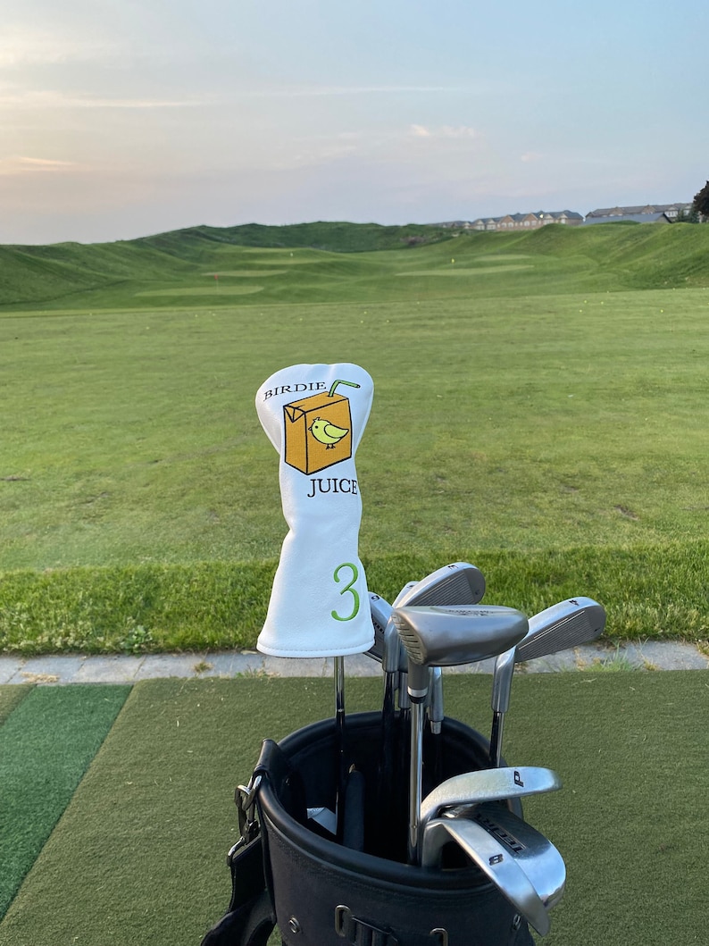 May include: A white golf club head cover with a cartoon illustration of a juice box with a bird on it. The text on the juice box reads "Birdie Juice". The club head cover has a green number "3" on it. The club head cover is on a golf club in a black golf bag with other golf clubs.
