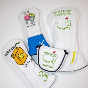 May include: Four white golf head covers with different designs. One cover has a cartoon ice cream cone and milkshake with the text "Shake or Shank". Another cover has a cartoon juice box with a bird and the text "Birdie Juice". The third cover has a green outline of the United States with a red flag and the text "Mulligan Masters". The fourth cover has a green outline of the United States with a red flag and the text "Three Putt Masters".