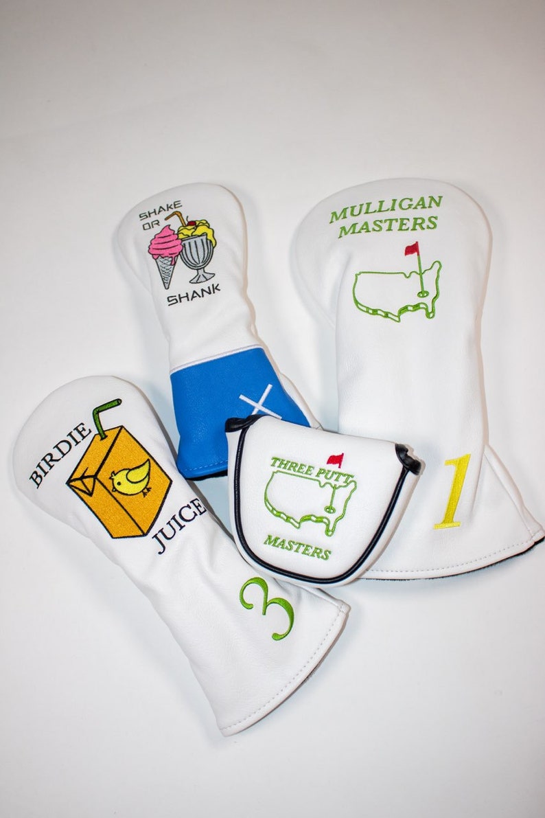 May include: Four white golf head covers with different designs. One cover has a cartoon ice cream cone and milkshake with the text "Shake or Shank". Another cover has a cartoon juice box with a bird and the text "Birdie Juice". The third cover has a green outline of the United States with a red flag and the text "Mulligan Masters". The fourth cover has a green outline of the United States with a red flag and the text "Three Putt Masters".