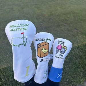 May include: Three white golf head covers with different designs. The first cover has the text "Mulligan Masters" and a green outline of the United States with a red flag. The second cover has the text "Birdie Juice" and a cartoon illustration of a juice box with a straw and a yellow bird. The third cover has the text "Shake or Shank" and a cartoon illustration of a milkshake with a cherry and whipped cream.