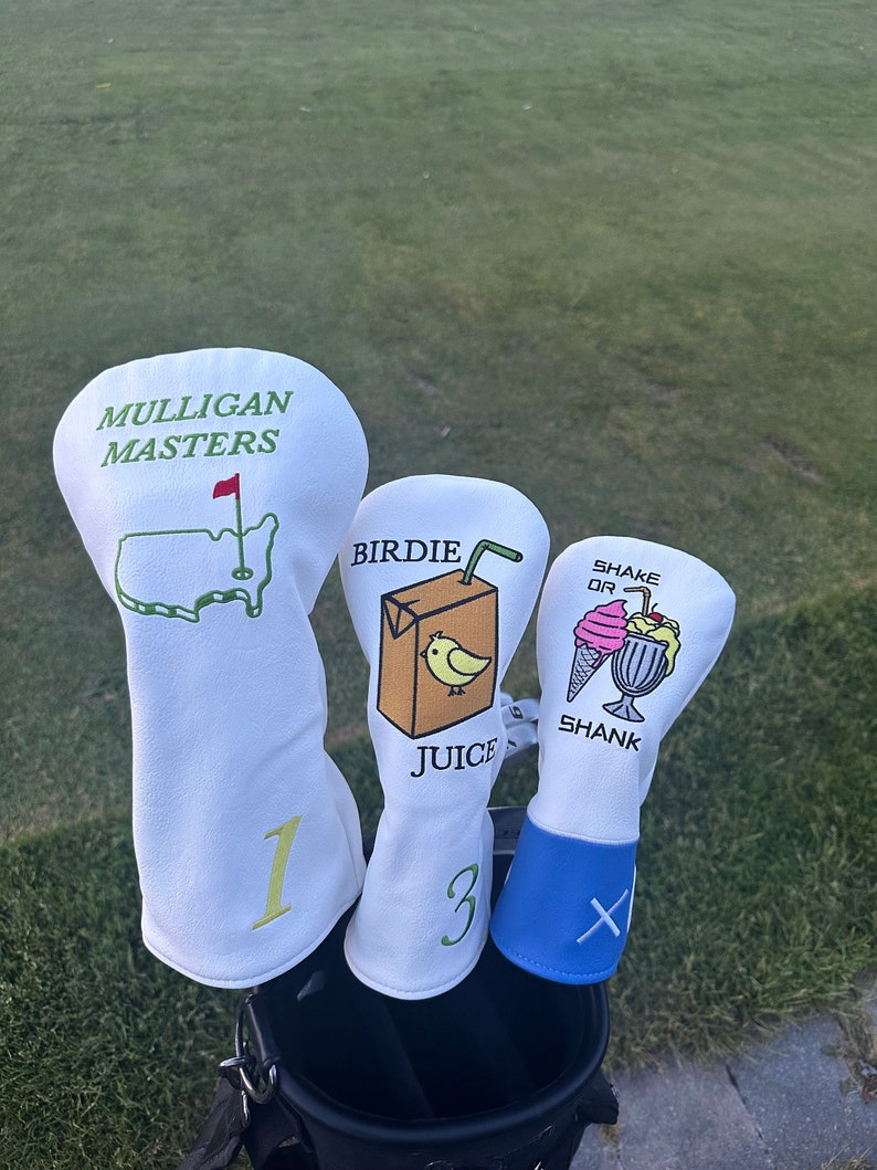 May include: Three white golf head covers with different designs. The first cover has the text "Mulligan Masters" and a green outline of the United States with a red flag. The second cover has the text "Birdie Juice" and a cartoon illustration of a juice box with a straw and a yellow bird. The third cover has the text "Shake or Shank" and a cartoon illustration of a milkshake with a cherry and whipped cream.
