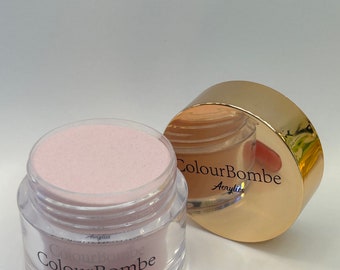 Puede incluir: Un frasco transparente con polvo acrílico rosa en su interior. El frasco tiene una tapa dorada con el texto "ColourBombe Acrylic". También se muestra una punta de uña acrílica rosa en la imagen.