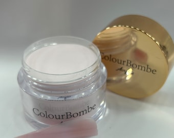 Puede incluir: Un tarro transparente con polvo rosa en su interior. El tarro tiene una tapa dorada con el texto "ColourBombe" en ella. Una punta de uña rosa está en primer plano.