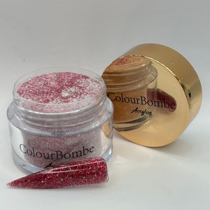 Puede incluir: Un frasco transparente lleno de purpurina roja y blanca, etiquetado como "ColourBombe Acrylics". El frasco está al lado de una tapa dorada con la misma etiqueta. Una punta de uña de purpurina roja y blanca está en primer plano.