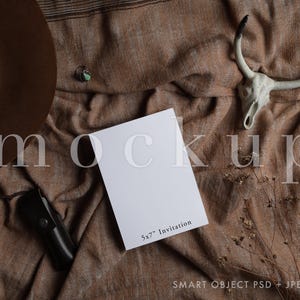 May include: A white 5x7 inch invitation card mockup on a brown fabric background with a cowboy hat, a small ring, and a white skull with horns.