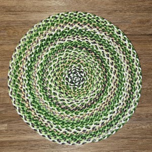May include: A circular braided mat featuring a variegated pattern of intertwined green, white, and dark brown fibers. The textile construction displays a consistent, tightly woven spiral design that radiates from the center to the outer edge.