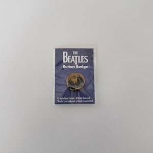 May include: A button badge featuring the Beatles. The badge is round with a photo of the band members. The badge is on a blue card with the band's name and the words "Button Badge".
