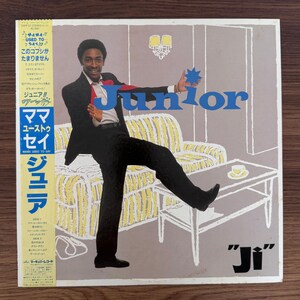 Pode incluir: Uma capa de disco de vinil com uma pessoa de terno preto e camisa branca posando em frente a uma ilustração de linhas de um sofá e uma mesa de centro. O texto "Junior" está impresso em letras azuis grandes e "Ji" em preto na parte inferior.
