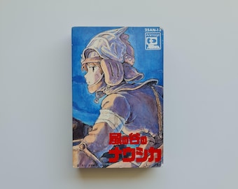 May include: A blue and white anime manga book cover featuring a character with a helmet and a blue background. The title of the book is "風の谷のナウシカ" in red Japanese characters.
