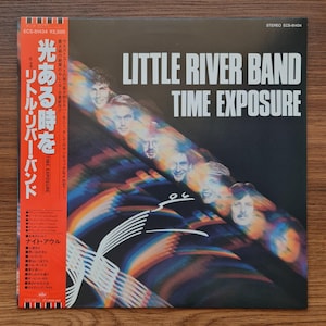 Puede incluir: Portada del álbum de vinilo "Time Exposure" de Little River Band. La portada presenta el nombre de la banda y el título del álbum en texto blanco sobre un fondo negro. También hay una imagen borrosa y colorida de los miembros de la banda.