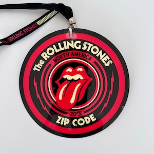 May include: A black and red circular The Rolling Stones North America 2015 Zip Code pass with the band's iconic tongue and lips logo. The pass is attached to a black lanyard with "Rolling Stones" printed on it.