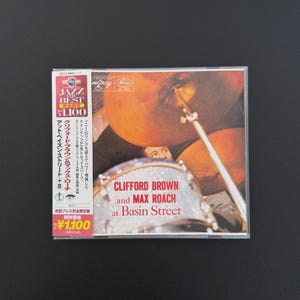 May include: A CD case for the album Clifford Brown and Max Roach at Basin Street. The cover features a close-up of a drum set with a prominent cymbal and drum head. A vertical white obi strip on the left side contains Japanese text and logos.