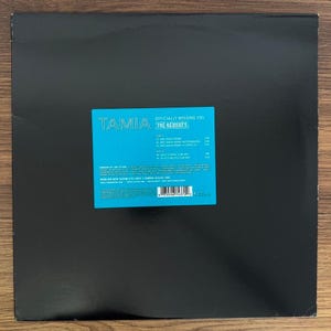 May include: A black vinyl record sleeve featuring a bright cyan rectangular sticker in the center. The sticker displays the text Tamia Officially Missing You The Remixes, along with a tracklist for Side A and Side B, production credits, and a barcode.