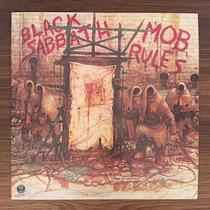 Könnte beinhalten: Ein quadratisches Vinyl-Schallplattencover mit dem Album-Artwork von Black Sabbath Mob Rules. Die Illustration zeigt vermummte Gestalten in zerlumpten Gewändern vor einer Holzkonstruktion mit einem blutbefleckten Tuch vor einer Ziegelmauer.