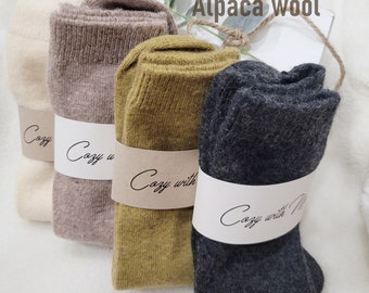 May include: Four pairs of alpaca wool socks in beige, mustard yellow, and grey. The socks are folded and stacked on top of each other. Each pair has a label that says "Cozy with Me".