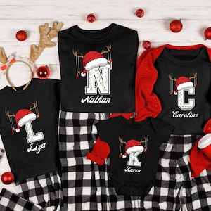 May include: A family of four wearing matching black Christmas pyjamas with a red Santa hat and reindeer antlers. The pyjamas feature a large letter with lights and the name of each family member. The pyjamas are on a white wood background.