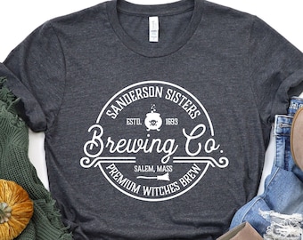 May include: A dark gray t-shirt with a white circle graphic that says "Sanderson Sisters Brewing Co. Est. 1693 Salem, Mass. Premium Witches Brew". The shirt is laid out on a surface with a green knitted sweater, two small orange pumpkins, and a pair of brown ankle boots.