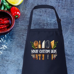 May include: A navy blue kitchen apron with a white design of kitchen utensils and the text "Your Custom Here".