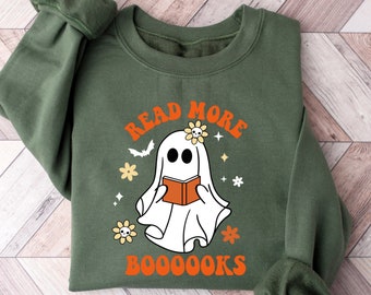 Puede incluir: Sudadera verde con un fantasma blanco leyendo un libro. El texto en la sudadera dice "Read More Booooks" con letras naranjas y un diseño floral.
