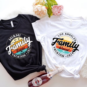 May include: Two black and white t-shirts with a retro-style graphic design. The graphic features a circle with the word "Family" in a stylized font, a sunset, and the text "Miami Vacation 2024" and "Los Angeles Vacation 2024".