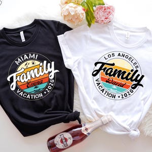 May include: Two black and white t-shirts with a retro-style graphic design. The graphic features a circle with the word "Family" in a stylized font, a sunset, and the text "Miami Vacation 2026" and "Los Angeles Vacation 2026".