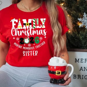 May include: A red t-shirt with the words "FAMILY CHRISTMAS 2020" in white, with festive holiday graphics. The shirt also says "MAKING MEMORIES TOGETHER SISTER". A Santa-themed mug is held in the hand.