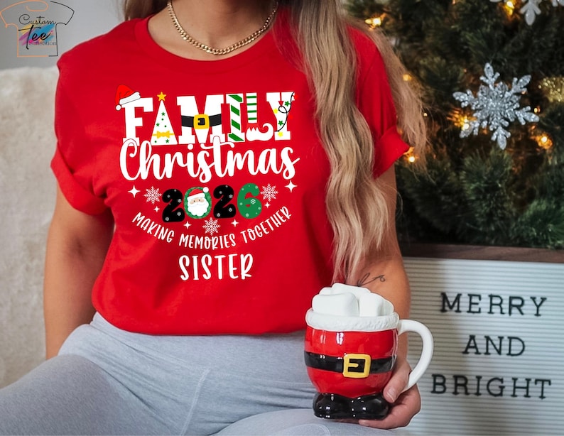 May include: A red t-shirt with the words "FAMILY CHRISTMAS 2020" in white, with festive holiday graphics. The shirt also says "MAKING MEMORIES TOGETHER SISTER". A Santa-themed mug is held in the hand.