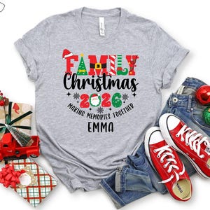 May include: A grey t-shirt with a Christmas theme. The shirt displays "FAMILY CHRISTMAS 2026" in red, green, and black, with "EMMA" underneath. Red trainers and denim jeans are also visible in the image, along with Christmas decorations.