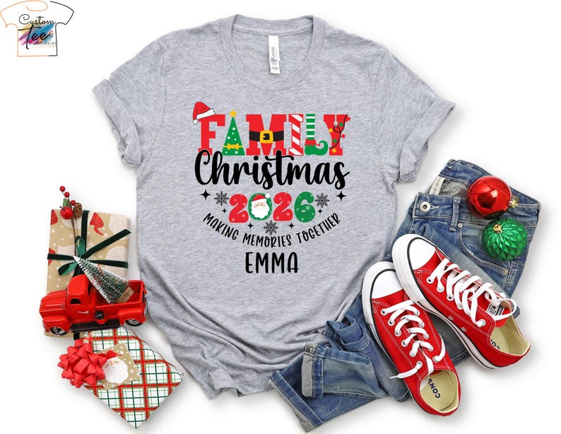 May include: A grey t-shirt with a Christmas theme. The shirt displays "FAMILY CHRISTMAS 2026" in red, green, and black, with "EMMA" underneath. Red trainers and denim jeans are also visible in the image, along with Christmas decorations.
