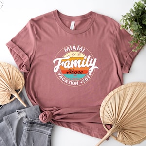 May include: A pink t-shirt with a vintage-style graphic that says "Miami Family Vacation 2026" with a palm tree and sun graphic. The text "Mama" is in a smaller font below the word "Family".