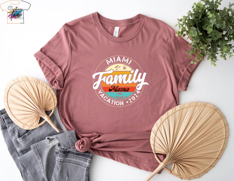 May include: A pink t-shirt with a vintage-style graphic that says "Miami Family Vacation 2026" with a palm tree and sun graphic. The text "Mama" is in a smaller font below the word "Family".