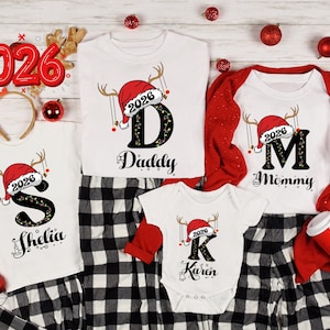 May include: A set of white t-shirts and a baby bodysuit featuring black letters with Christmas light patterns, topped with red Santa hats and reindeer antlers. The designs include the year 2026 and names including Daddy, Mommy, Shelia, and Karen.