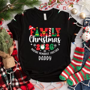 May include: Black t-shirt with the text "Family Christmas 2026 Making Memories Together Daddy" in red, green, and white. The shirt has a Christmas tree and Santa hat graphic. The shirt is displayed with Christmas decorations.