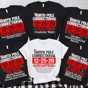 May include: A collection of five t-shirts, four black and one white, featuring the text North Pole Correctional, the date 12-25-26, and various humorous criminal charges like Felony Assault, Identity Theft, Disturbing the Peace, and Kidnapping.
