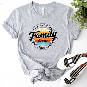 May include: Gray t-shirt with a circular design featuring the text "Los Angeles Family Vacation 2026" with a sunset graphic and the name "Emma" in the center.