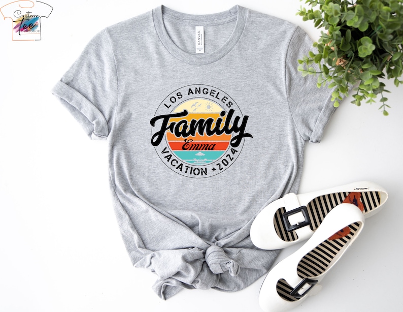 May include: Gray t-shirt with a circular design featuring the text "Los Angeles Family Vacation 2026" with a sunset graphic and the name "Emma" in the center.