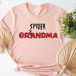 Puede incluir: Una camiseta rosa con el texto "SPIDER GRANDMA" en negro con un peque&ntilde;o gr&aacute;fico de Spiderman rojo y azul en el medio del texto.