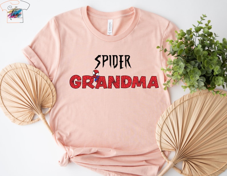 Puede incluir: Una camiseta rosa con el texto "SPIDER GRANDMA" en negro con un peque&ntilde;o gr&aacute;fico de Spiderman rojo y azul en el medio del texto.