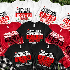 May include: A collection of white, red, and black t-shirts featuring the text North Pole Correctional, the date 12-25-26, and various humorous holiday-themed offenses like Arson, Theft, Fraud, and Disturbing the Peace, each accented with a small Santa hat.