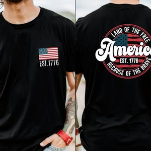 May include: A gray t-shirt with a small American flag on the front and a larger circle design on the back. The circle design features a larger American flag, the text "Land of the Free", "America", "Est. 1776", and "Because of the Brave".