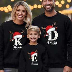 May include: Three people wearing matching black sweatshirts with a Christmas theme. Each sweatshirt features a letter, a Santa hat, and antlers. The names "Karen", "David", and "Ethan" are printed below the letters.