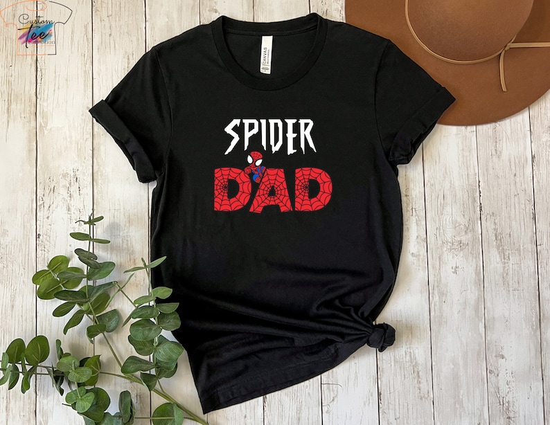 Puede incluir: Camiseta negra con un gr&aacute;fico blanco que dice "Spider Dad" con una imagen de dibujos animados de Spiderman en el medio de la palabra "Dad".