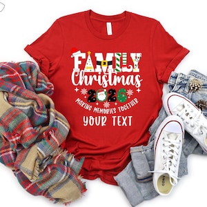 May include: A red t-shirt featuring a Christmas design with the text "Family Christmas 2020". The graphic includes festive symbols such as a Santa hat and Christmas tree. The shirt is styled with jeans, white trainers, and a tartan scarf.