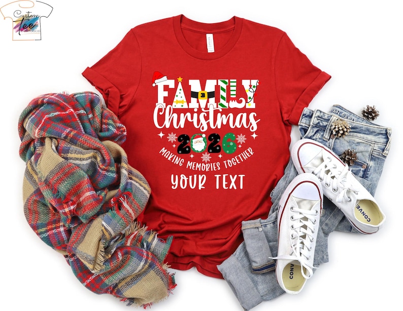 May include: A red t-shirt featuring a Christmas design with the text "Family Christmas 2020". The graphic includes festive symbols such as a Santa hat and Christmas tree. The shirt is styled with jeans, white trainers, and a tartan scarf.