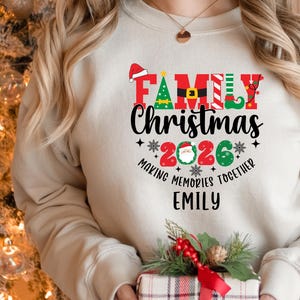 May include: A cream-coloured sweatshirt featuring a "Family Christmas 2026" design. The design includes a Santa hat, Christmas tree, and elf shoe. The text "Making Memories Together" and a name are also featured. A gift box with a red bow is in the foreground.